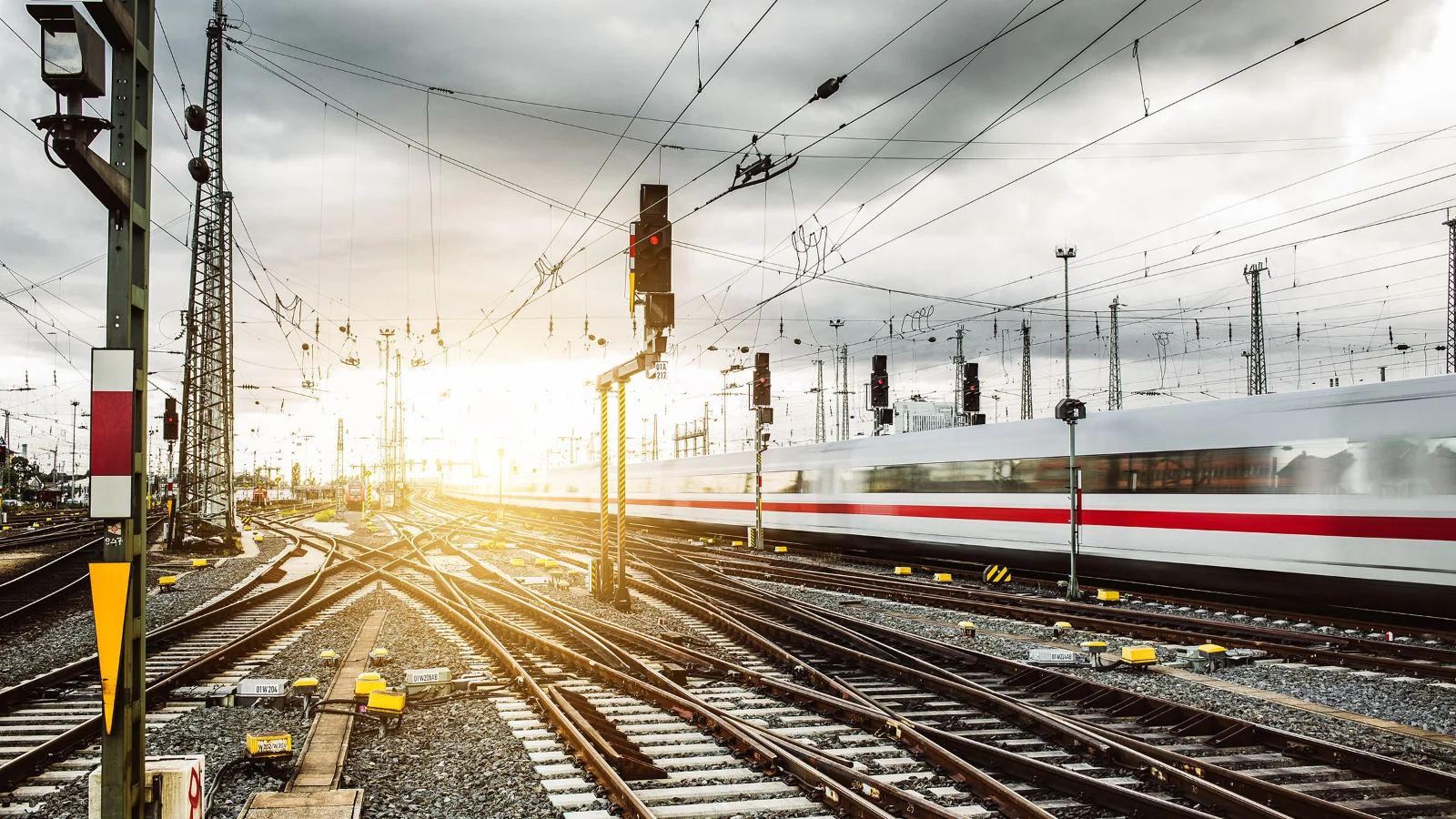 Vector supports evolving rail systems with development and validation workflows that improve long-term operational reliability. Image of multiple railway tracks, overhead power lines, and a fast passenger train moving through a bright rail yard at sunrise.